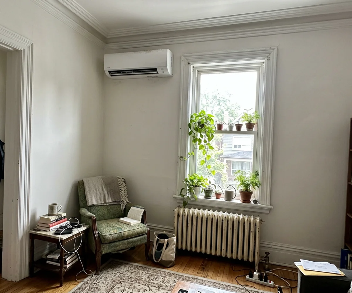 Ductless heat pump wall head above a window in a Victorian Toronto living room with the original radiator below